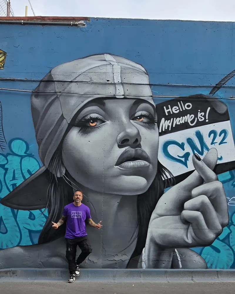 Murale fotorealistico di volto femminile in black and grey con cartello “Hello my name is” su parete blu